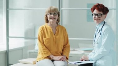 Pozitif kıdemli kadın ve genç bayan doktor klinikteki tıbbi kanepede oturup kameraya bakıp gülümsüyor.
