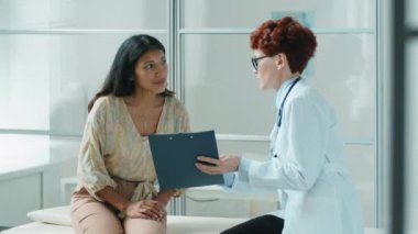 İspanyol kadın panoya onay formu imzalıyor ve klinikteki kanepede birlikte otururken beyaz kadın doktorla konuşuyor.