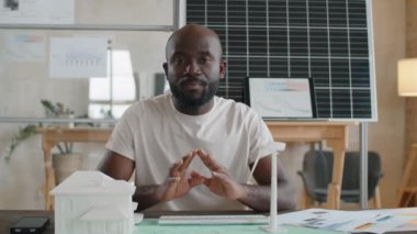 Ofiste oturan Afro-Amerikan mühendis, kamerada kağıt planı modellerini, yel değirmeni türbini ve ev modelini gösteriyor ve çevrimiçi internet üzerinden yenilenebilir enerji projesini anlatıyor.