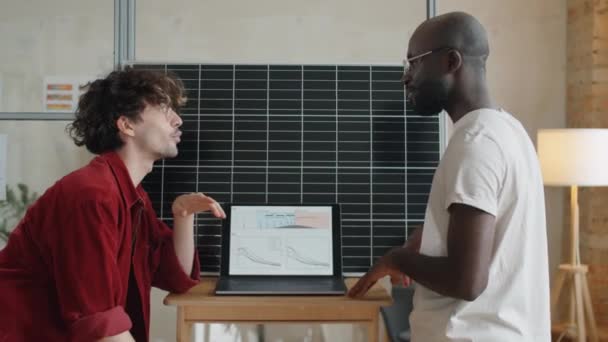 アフリカ系アメリカ人と白人男性の研究者らは オフィスの太陽光パネルの横に立ちながら ノートパソコン画面上で再生可能エネルギープロジェクトについて議論した — ストック動画