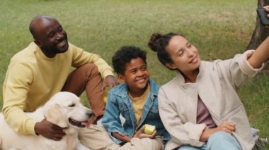 Neşeli Afro-Amerikan ailesi Golden Retriever köpeğiyle parkta oturuyor ve yaz pikniği yaparken akıllı telefonla selfie çekiyor.