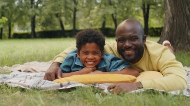 Mutlu Afro-Amerikan baba ve küçük oğlunun portresi parkta battaniyeye uzanmış kameraya bakıyor ve gülümsüyor.