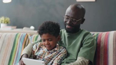 Neşeli Afro-Amerikalı çocuk ve babası evdeki kanepede oturmuş, gülümsüyor ve dijital tablette oynuyorlar.