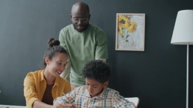 Küçük Afrikalı çocuk evde ebeveynlerinin yardımıyla ödevlerini yaparken mutlu anne ve babayla konuşurken deftere not alıyor.
