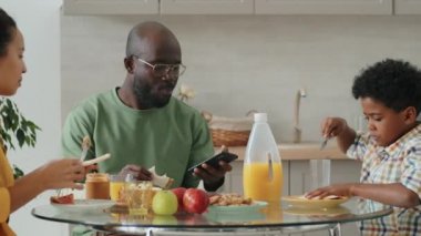 Afro-Amerikalı bir adam evde karısı ve oğluyla kahvaltı ederken akıllı telefon kullanıyor.