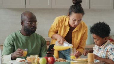 Küçük Afro-Amerikalı çocuk tostun üstüne fıstık ezmesi koyuyor ve kahvaltıda anne ve babasıyla konuşuyor.