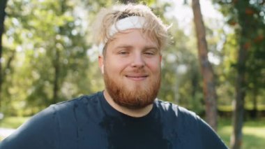 Terli tişörtlü, kablosuz kulaklıklı, parkta durup antrenmandan sonra kameraya bakıp gülümseyen pozitif kilolu adamın göğüs portresi.