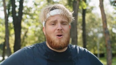 Terli tişörtlü yorgun bir adamın göğüs portresi. Parkta durmuş kameraya bakıyor ve koştuktan sonra derin nefes alıyor.