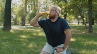 Spor giyimli beyaz bir adamın parkta mindere oturup antrenmandan sonra şişeden taze detoks smoothie içerkenki pozunu çek.