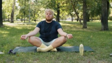 Nilüferde oturan şişman genç adam gözleri kapalı, elleri çamurlu parkta meditasyon yaparken yoga minderinde poz veriyor.