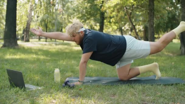太りすぎの男は ラップトップ上でオンラインヨガのトレーニングを見て 公園でマット上で反対側の腕や脚の拡張を練習 — ストック動画