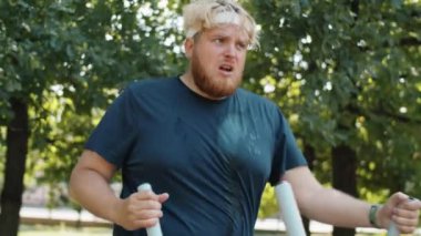 Aşırı kilolu bir adamın terli spor kıyafetleriyle parkta yürüyen bir makine üzerinde egzersiz yapmasını.