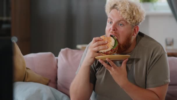 Толстый Человек Ест Вкусный Гамбургер Смотрит Телевизор Сидя Диване Гостиной — стоковое видео