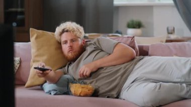 Şişman adam evdeki kanepede uzanıp cips yiyor ve uzaktan kumandayla televizyon kanallarını değiştiriyor.