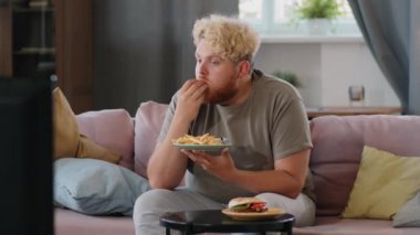 Evdeki koltukta oturan, ketçaplı patates kızartması yiyen ve televizyon izleyen obez bir genç.