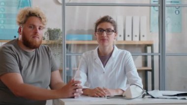 Obeziteli genç bir kadın doktor ve adamın, muayenehanedeki masasında oturup, kamera önünde poz verirken çekilen görüntüler.