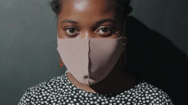 Siyah arka planda poz veren koruyucu maskeli Afro-Amerikalı genç bir kadının portre fotoğrafı.