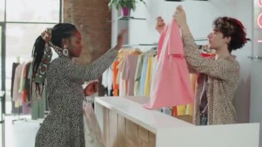 Kafkasyalı genç satış asistanı elbise mağazasında çalışırken Afro-Amerikalı kadın müşteriler için kağıt torba içinde tişört topluyor.