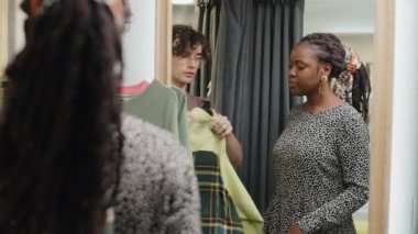 Beyaz tenli tezgahtar, genç Afro-Amerikalı kadına soyunma odasında yeni kıyafetler seçmesinde yardım ediyor.