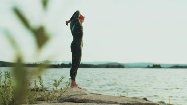 岩の多い湖畔に立っている水着で女性のトライアスロンのショットを傾け 彼女の筋肉を伸ばす前に水泳 — ストック動画