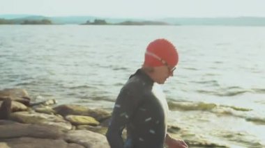 Mayo, kep ve gözlüklü kadın triatlet kayalık sahilde göle yürüyor ve yüzmeye hazırlanıyor.