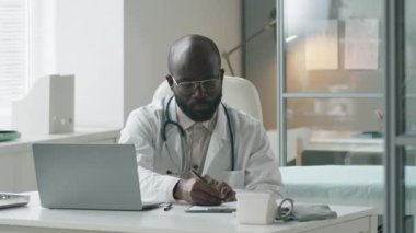 Boynunda steteskop olan Afrikalı Amerikalı bir erkek doktor klinikte masa başında dizüstü bilgisayar kullanıyor ve iş saatleri boyunca not alıyor.