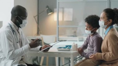Koruyucu maske ve beyaz önlüklü Afrikalı Amerikalı çocuk doktoru covid-19 salgınında klinikte küçük çocuk ve annesiyle konuşurken not alıyordu.