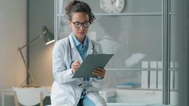 Beyaz önlüklü ve gözlüklü genç melez kadın doktor klinikteki kanepede oturuyor ve not alıyor.