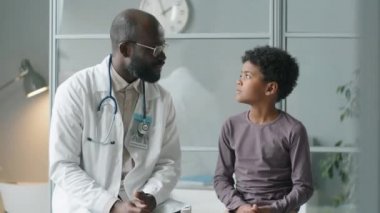 Afro-Amerikalı doktor ve küçük çocuk klinikteki tıbbi kanepede birlikte oturuyorlar, konuşuyorlar ve sonra kameraya bakıp gülümsüyorlar.