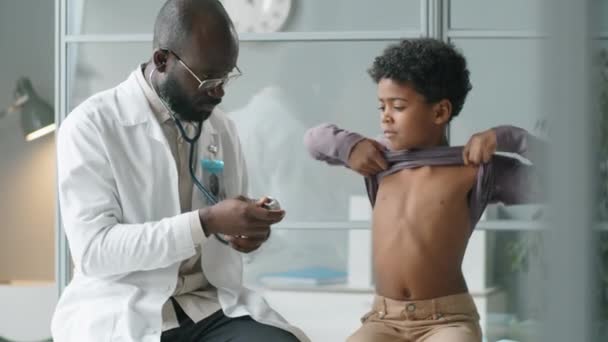 Врач Афроамериканец Белом Халате Используя Стетоскоп Слушает Сердцебиение Маленького Мальчика — стоковое видео
