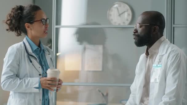 アフリカ系アメリカ人の男性医師がコーヒーを飲み 診療所での勤務中に休憩中に混合人種の女性同僚と話す — ストック動画