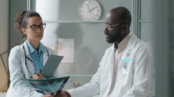 人種混合女性医師と彼女のアフリカ系アメリカ人男性の同僚は 診療所で一緒に作業しながら胸X線画像を議論 — ストック動画