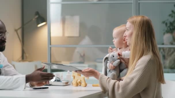 白人女性は赤ん坊の娘を抱えており 黒人小児科医に医学史を与え 診療所で相談中に彼と話をする — ストック動画
