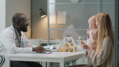 Neşeli Afrikalı Amerikalı pediyatrist notlar alıyor ve danışmanlık sırasında küçük kızını kollarına alan beyaz kadınla konuşuyor.