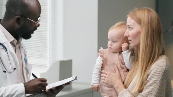 白人女性は赤ん坊の娘を抱えており アフリカ系アメリカ人男性医師と診療中にメモを書きながら話す — ストック動画