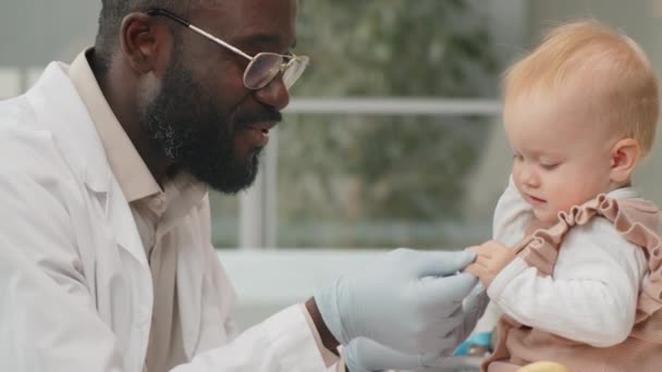 ポジティブアフリカ系アメリカ人小児科医で中にコートと保護手袋保持手の可愛いです1歳白人の赤ん坊ながら彼女の健康診断でクリニック — ストック動画