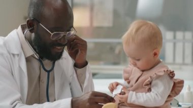 Afrikalı Amerikalı pediyatrist gülümseyip steteskop kullanırken klinikteki güzel beyaz bebeğe sağlık kontrolü yapıyor.