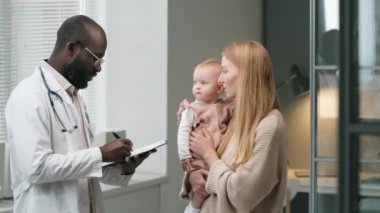 Afro-Amerikalı doktor not alıyor ve klinikte danışmanlık yaparken küçük kızını kollarına alan beyaz kadınla konuşuyor.