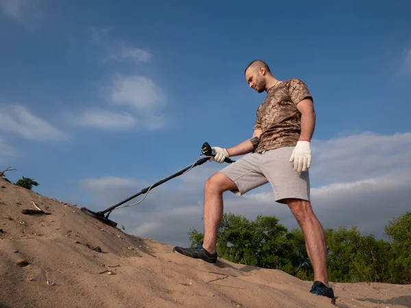 Man on a sandy ground with a metal detector looking for treasure on a sunny summer day.