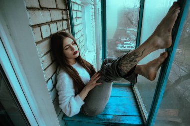 Balkonda poz veren güzel, genç ve seksi bir kız. Modern tarzda bir kızın resim portresi. Oturmuş ve fotoğrafa ses eklemiş..