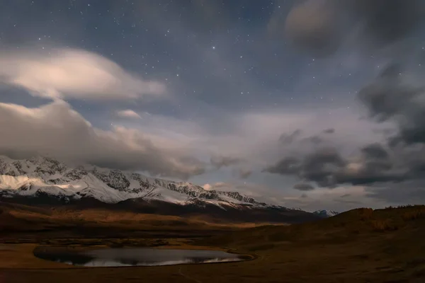 Amazing night landscape with snowy mountains, lake, beautiful storm clouds and stars in the sky in the moonlight