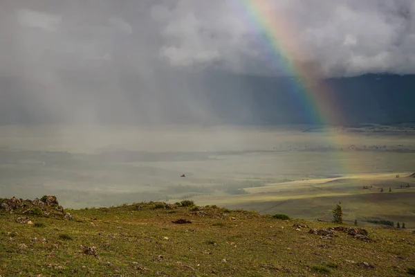 Amazing bright colorful rainbow over the mountains, steppe and valley against the backdrop of a stormy sky, clouds and rain. Altai, Russia.