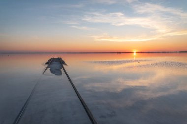 Güzel bulutlar, güneş, ufukta tren, suda yansımalar ve ön planda raylar olan tuzlu bir gölün üzerinde inanılmaz bir gün batımı. Bursol Gölü, Altai, Rusya.
