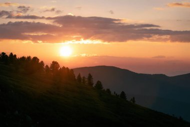 İnanılmaz turuncu günbatımı, dağların üzerinde yuvarlak bir güneş diski, dağların yamaçlarında sedir ormanı ve güneş ışığında gökyüzünde bulutlar.