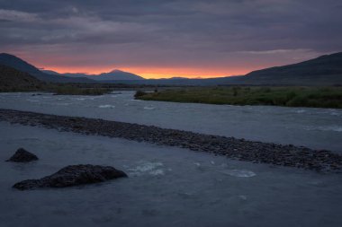 Dağlı turuncu gün doğumu, hızlı dağ nehri ve gökyüzünde fırtına bulutları. Altai, Rusya
