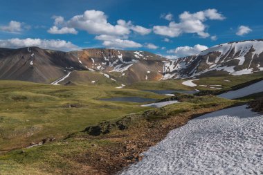 Dağlar arasındaki vadideki iki alp gölünün muhteşem akşam üstü manzarası, yamaçlarda kar, mavi gökyüzü ve bulutların arka planında toprak bir yol ve arabalar. Altai, Rusya