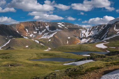 Dağlar arasındaki vadideki iki alp gölünün muhteşem akşam üstü manzarası, yamaçlarda kar, mavi gökyüzü ve bulutların arka planında toprak bir yol ve arabalar. Altai, Rusya