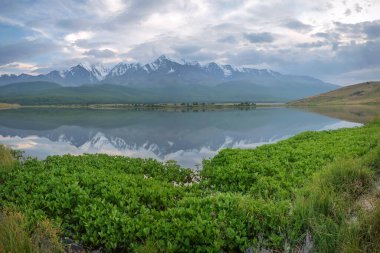 Karla kaplı dağlarla, ormanla, gölle ve göldeki güzel yansımalarla dolu muhteşem yaz manzarası bulutların ve mavi gökyüzünün arkaplanına karşı. Altai, Rusya