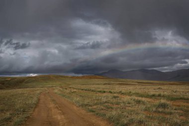 Karanlık ve fırtınalı bir gökyüzünde, dağların üzerinde yağmur ve bozkırda toprak bir yol olan inanılmaz bir gökkuşağı. Altai, Rusya
