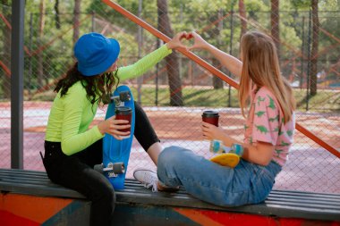 Modern bir kameranın önünde, iki güzel bayanı park edin ve kahve içerken eğlenin ve ellerden küçük şirin bir kalp yapın.